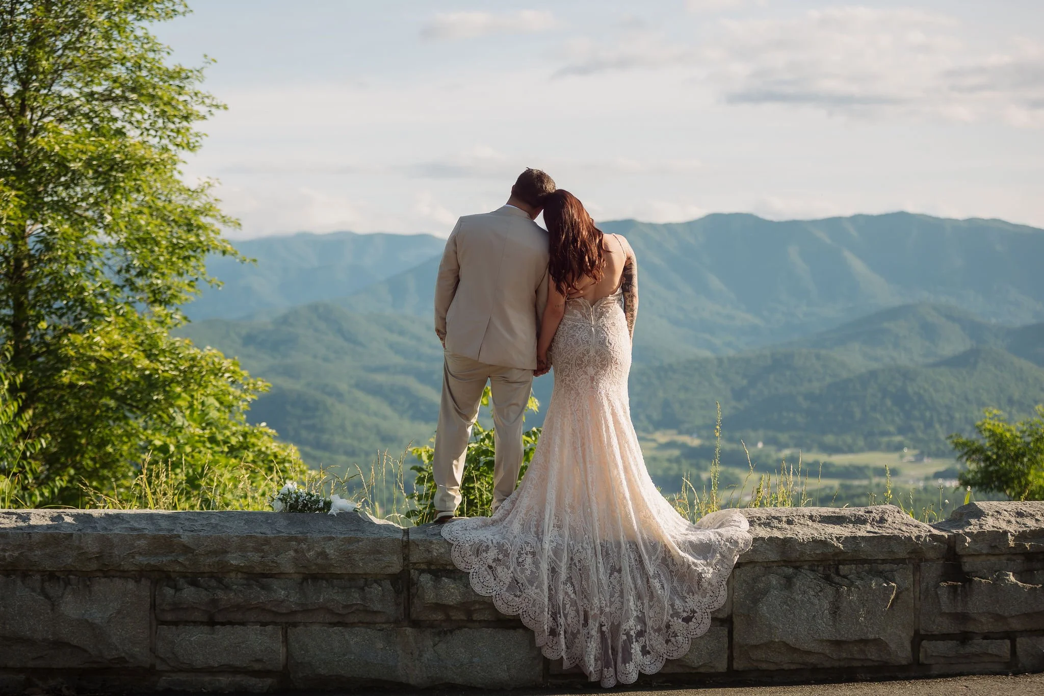 A couple visiting the Smokies shares a quiet moment overlooking layers of blue mountain ridges, with the bride's flowing lace gown draped over a stone wall along Foothills Parkway.
