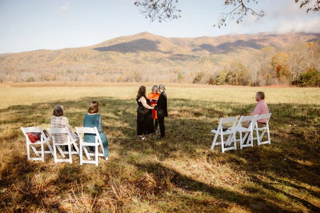 Wide shot of intimate Halloween Wedding ceremony with seated guests in open field, Gatlinburg photographer capturing small outdoor fall elopement with mountain views, autumn colors, and rustic countryside setting