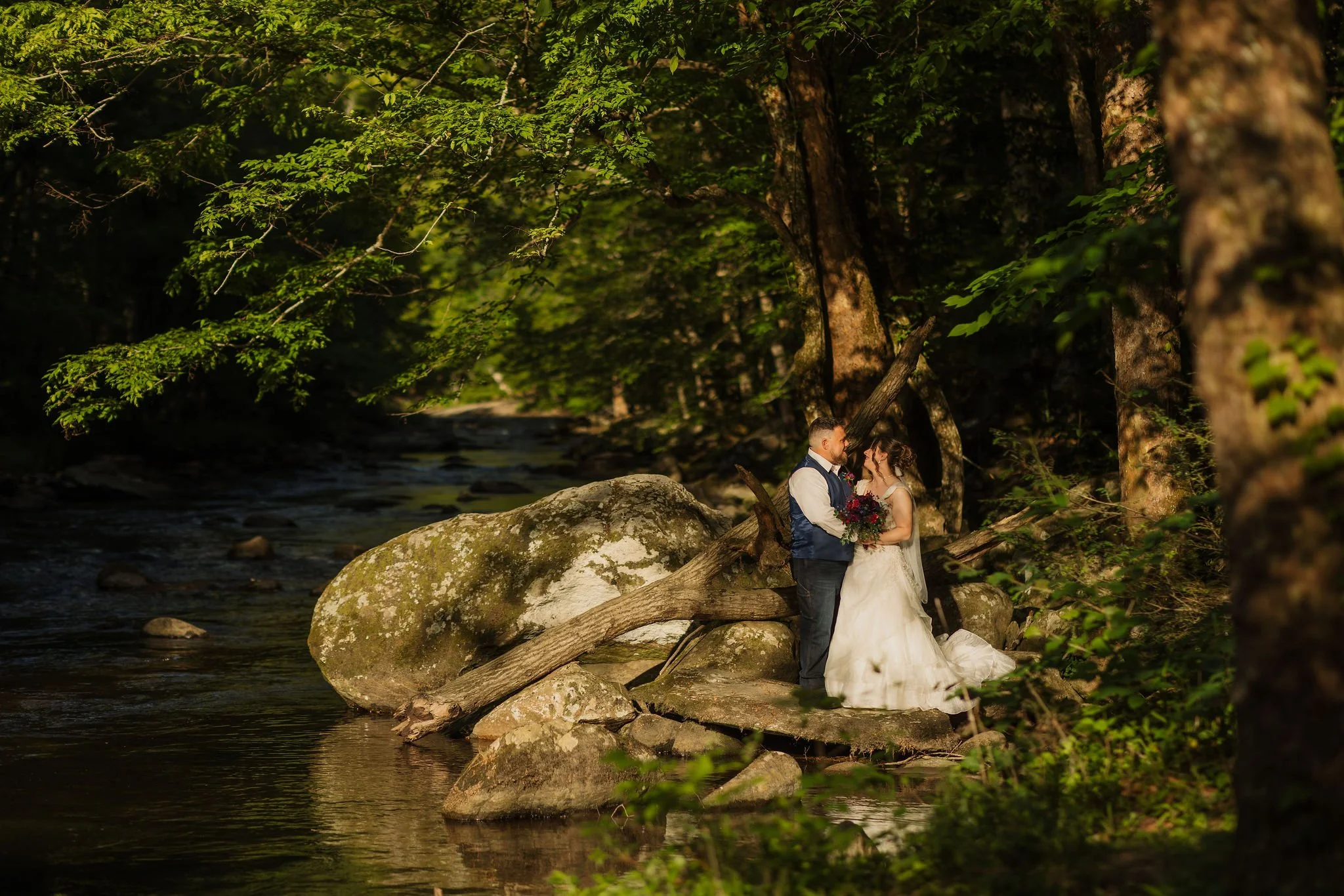 A bride and groom share an intimate moment on large moss-covered rocks beside a peaceful woodland creek, surrounded by lush green trees and golden sunlight during their Smoky Mountain elopement.