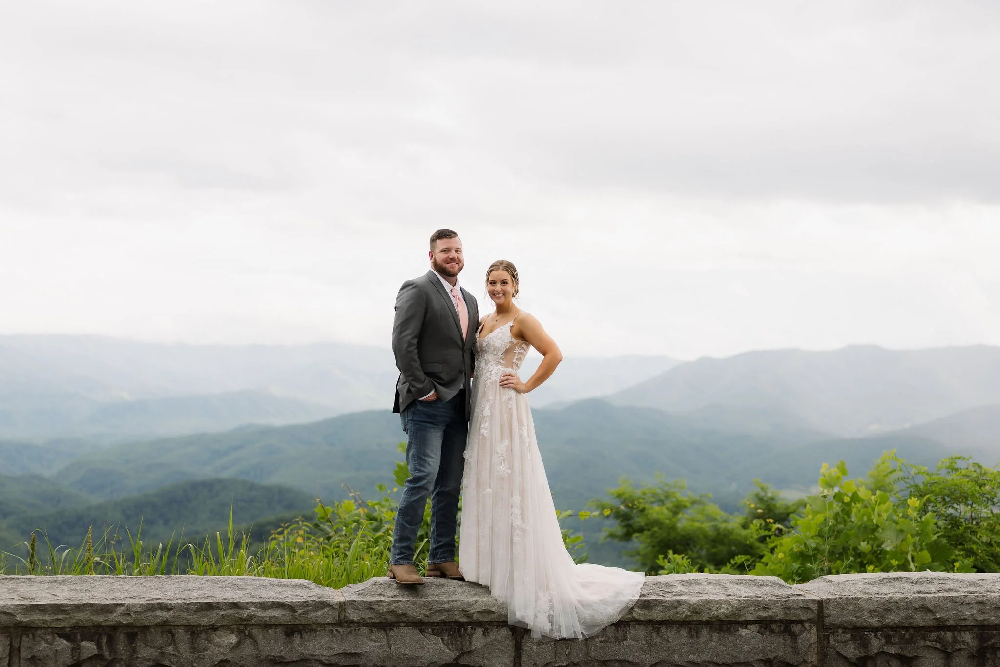 Bride and groom pose on a stone overlook wall with sweeping views of the Smoky Mountain foothills behind them during their intimate spring wedding near Gatlinburg.