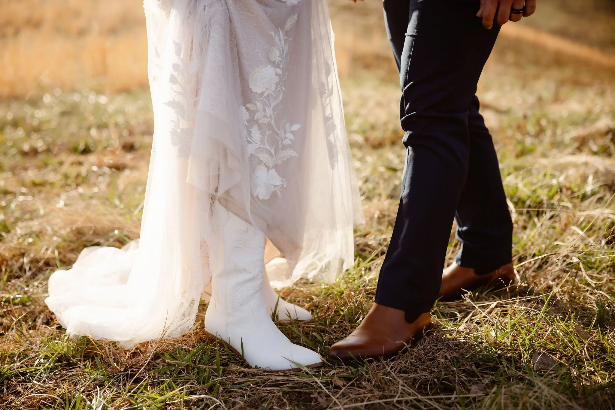 Close-up of bride and groom footwear in a sunlit field, featuring white cowboy boots beneath a lace wedding dress during wedding traditions while eloping to Gatlinburg with a Gatlinburg photographer.