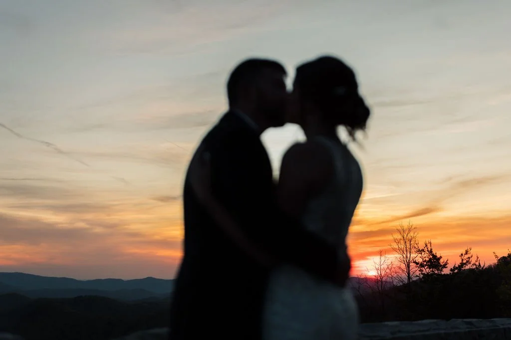 A softly blurred silhouette of a bride and groom sharing a kiss is set against a vibrant sunset over the Smoky Mountains.
