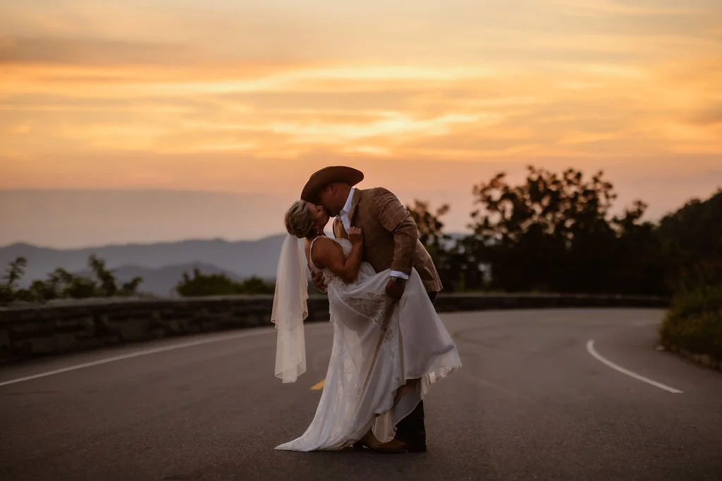Bride and groom sharing a romantic dip and kiss in the middle of Foothills Parkway during their sunset elopement, with a glowing Smoky Mountain sky and layered mountain views creating a dramatic backdrop for their Smoky Mountain elopement.