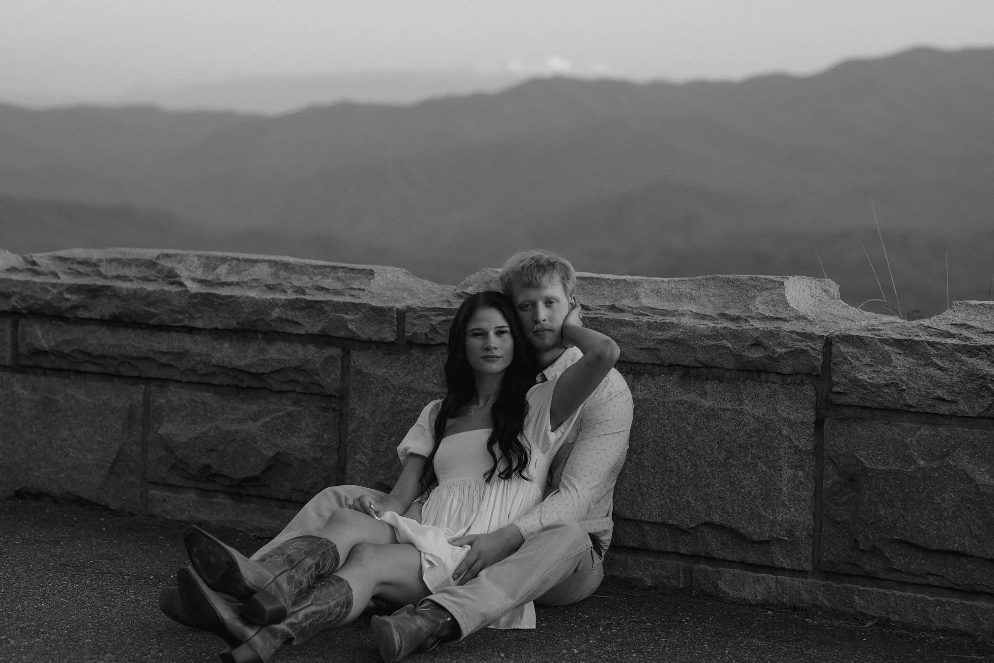 Couple sitting together against a stone overlook wall in a black-and-white portrait, embracing while overlooking layered mountain views in the background.