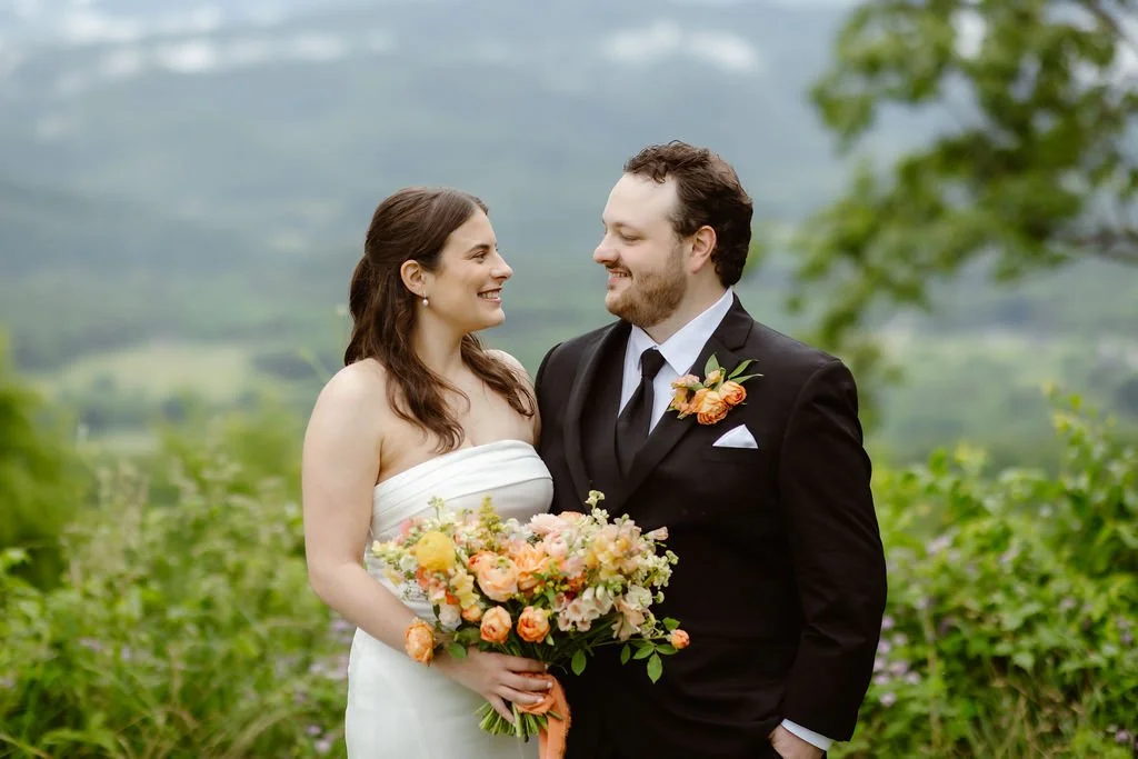 Bride and groom smiling at each other while standing in front of a misty mountain landscape, with the bride holding a pastel spring bouquet—an intimate portrait from Smoky Mountain Elopements in the Spring, photographed by a Gatlinburg photographer.