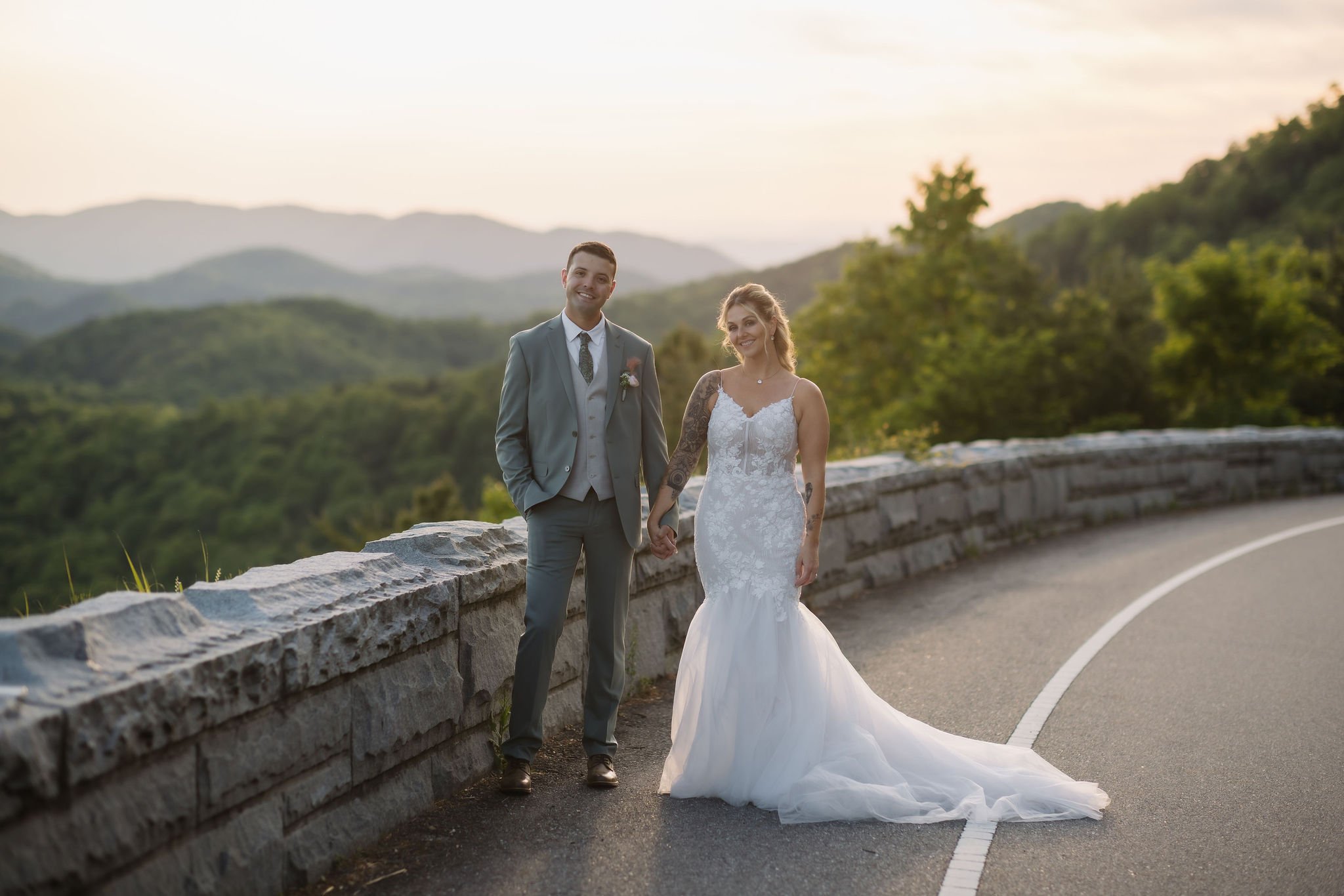 Newlyweds hold hands beside the stone wall and winding road of Foothills Parkway, surrounded by layered mountain views at sunset.