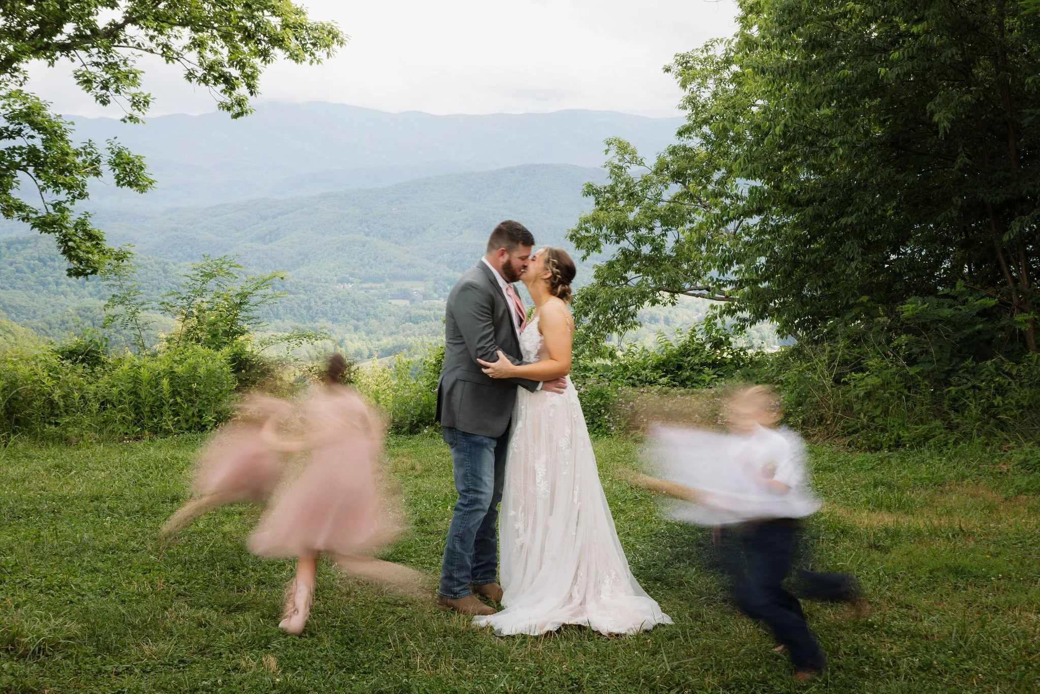 Bride and groom share a kiss during their Gatlinburg elopement as children play around them, with lush greenery and sweeping views of the Smoky Mountain foothills in the background.