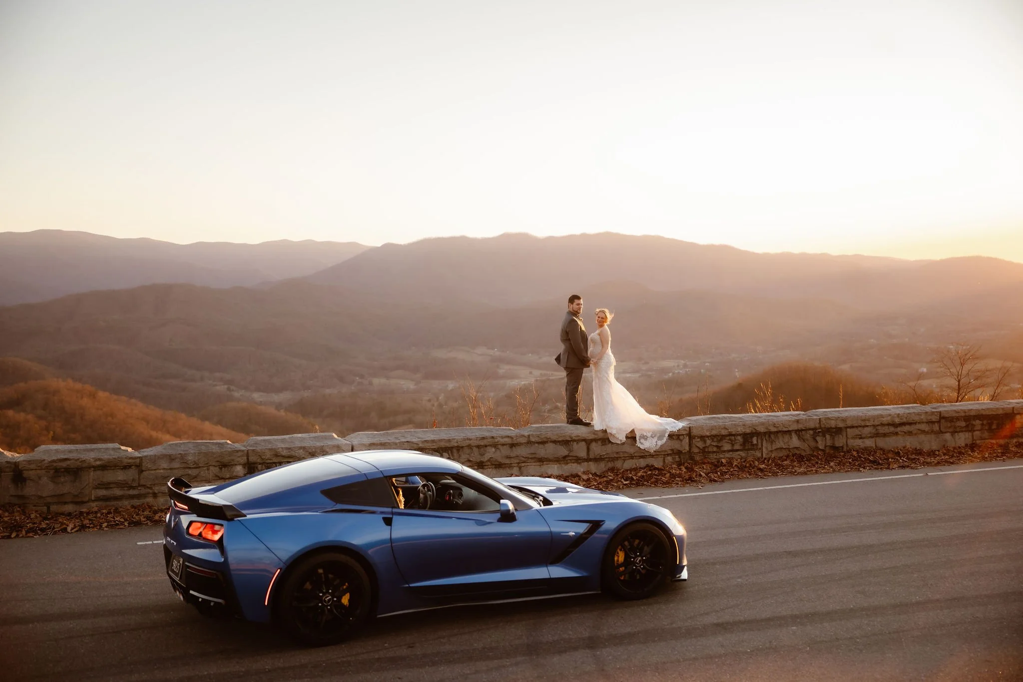 Bride and groom posing beside a blue sports car at a scenic mountain overlook during sunset, captured by a Gatlinburg wedding photographer for pregnant wedding pictures.
