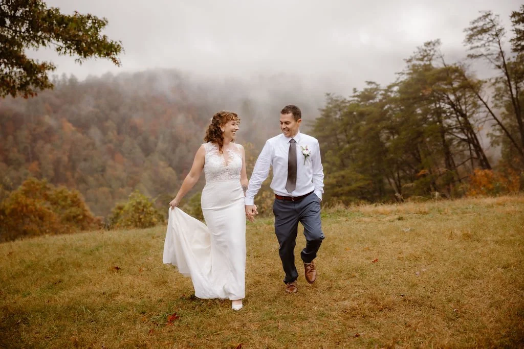 Bride and groom walking hand in hand across misty mountain field during rain on my wedding day Foothills Parkway elopement, romantic outdoor ceremony moment captured by Gatlinburg photographer with soft fog and autumn landscape