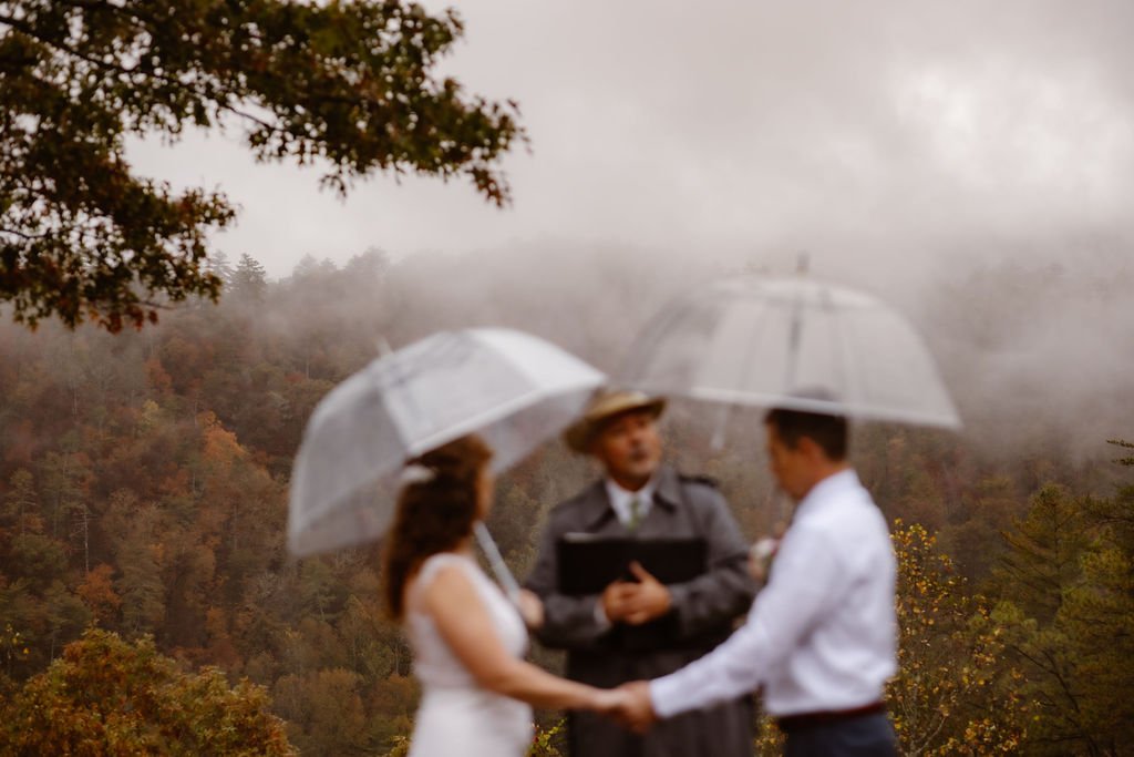 Couple exchanging vows under clear umbrellas during rain on my wedding day Foothills Parkway elopement, captured by Gatlinburg photographer with misty Smoky Mountains scenery, autumn foliage, and intimate rainy mountain wedding ceremony
