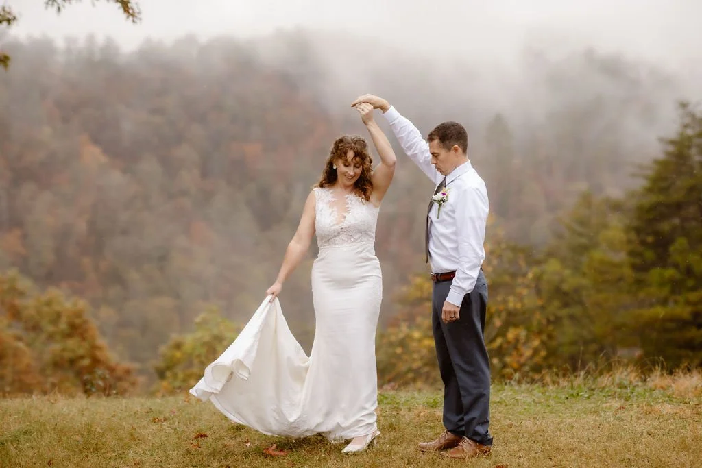 Bride twirling in wedding dress while groom holds her hand during rain on my wedding day Foothills Parkway elopement, playful candid moment captured by Gatlinburg photographer with misty mountain and fall foliage backdrop