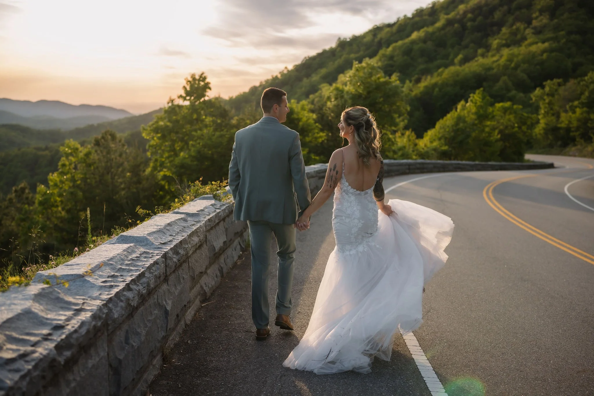 Newlyweds walk hand in hand along Foothills Parkway at sunset, with a winding mountain road and forested Smoky Mountains behind them.