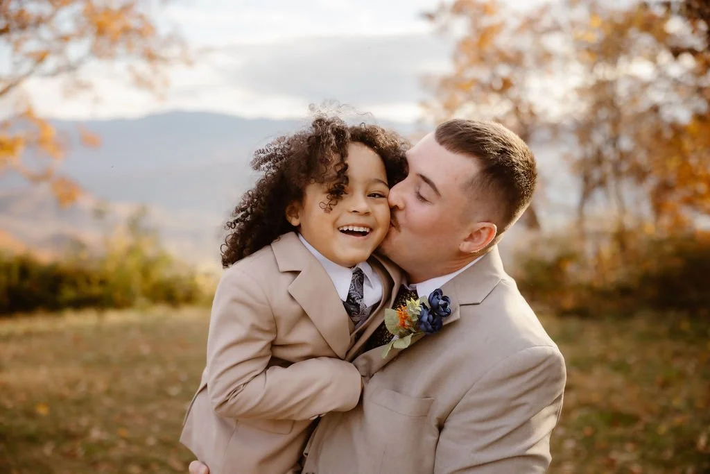 Groom kissing child’s cheek during outdoor wedding portrait with autumn mountains in background, foothills parkway elopement photography