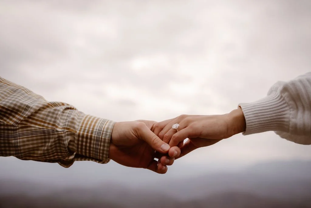 Close-up of couple holding hands with engagement ring centered against Foothills Parkway mountain backdrop, styled engagement photos clothes in neutral tones captured by Gatlinburg photographer during romantic proposal session