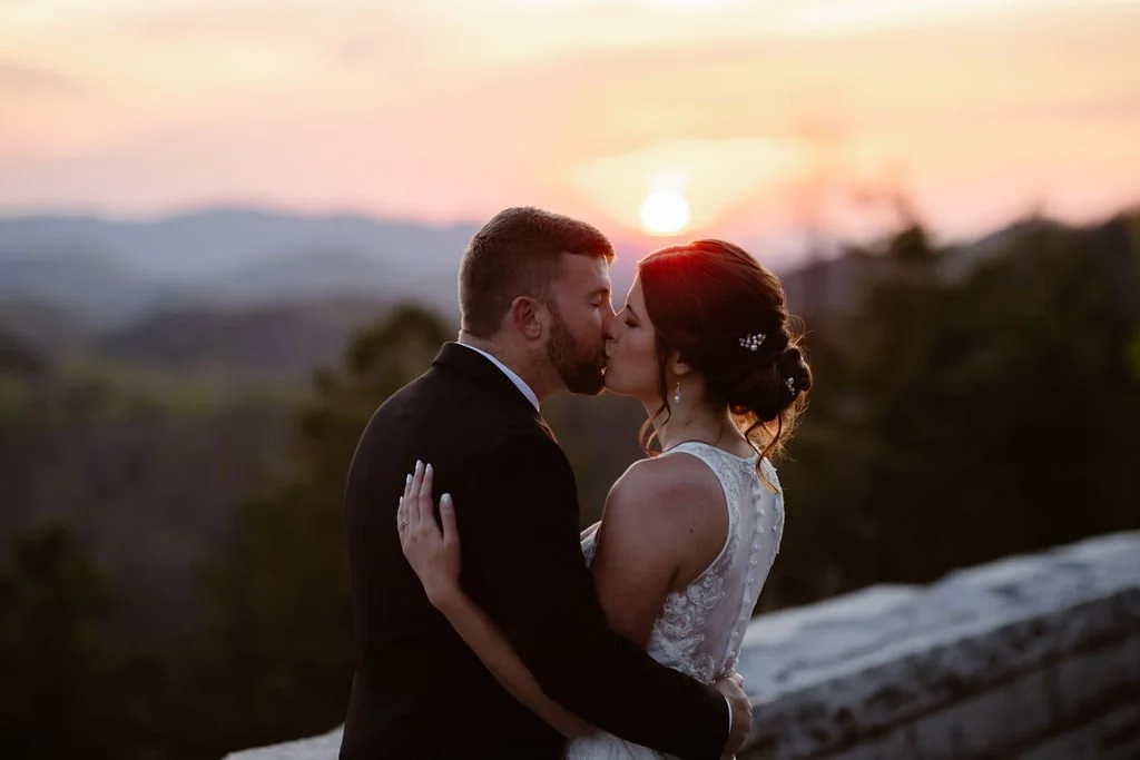 A bride and groom share a kiss at sunset on the Foothills Parkway, with the glowing sun dipping behind the Smoky Mountains in the distance