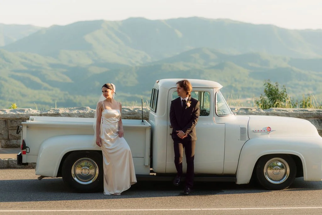 Bride and groom pose beside a vintage white pickup truck on the foothills parkway, with sweeping Smoky Mountain foothills filling the background.
