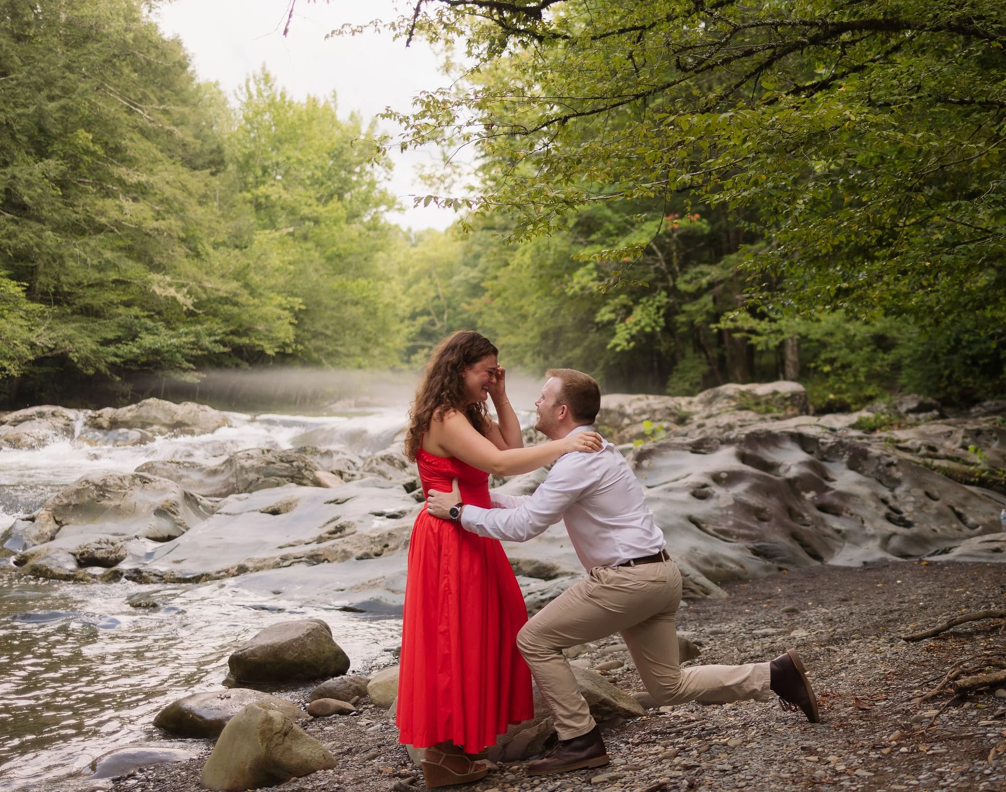 Man drops to one knee for a surprise proposal beside a gatlinburg waterfall in greenbrier as his partner reacts.