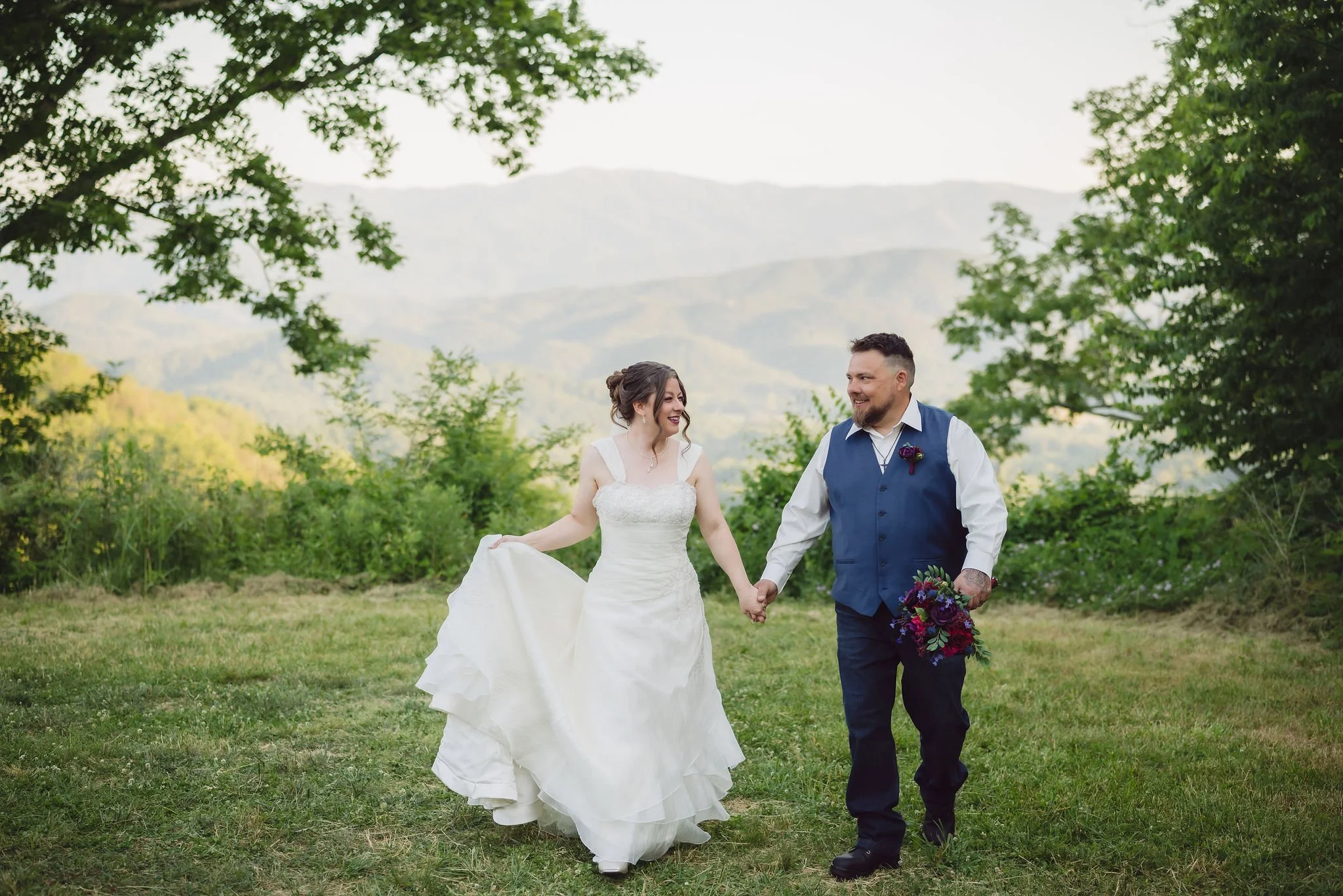 A bride and groom walk hand in hand through a grassy mountain meadow, smiling at each other with scenic Smoky Mountain ridges behind them during their Gatlinburg elopement.