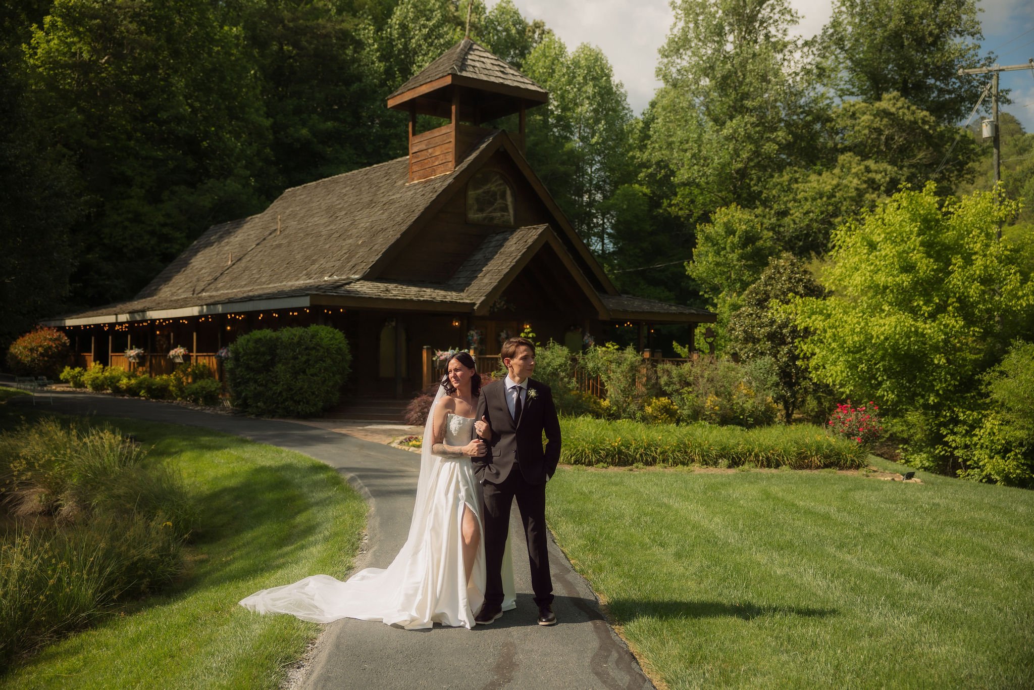 Newlyweds pose together on a winding path in front of the rustic Little Log Chapel in Gatlinburg, surrounded by green lawns and trees.