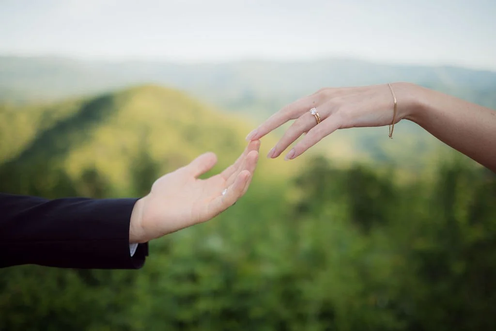 Close-up of the newlyweds reaching toward each other, highlighting the bride’s engagement ring against the mountain scenery along the foothills parkway.
