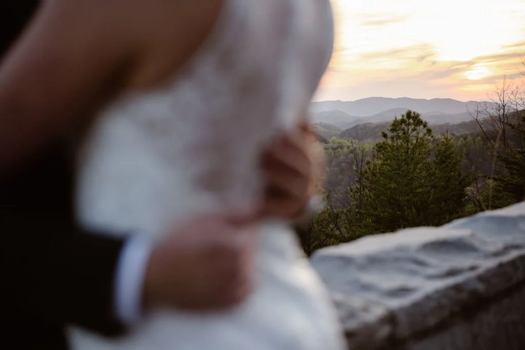 A softly focused embrace between a bride and groom frames a glowing sunset over the Smoky Mountains from the Foothills Parkway