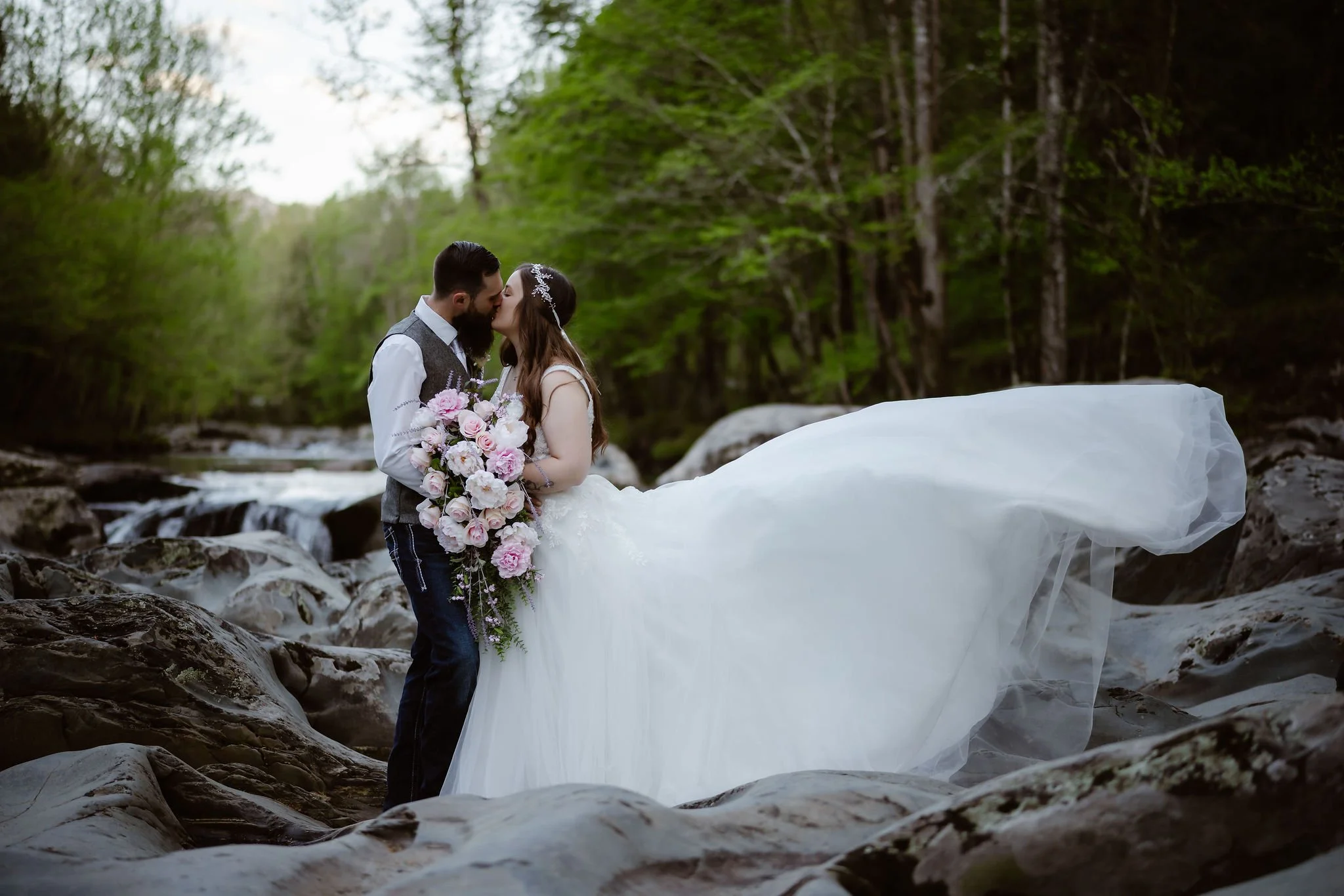 Elopement photography at Greenbrier in the Smoky Mountains featuring a bride and groom sharing a kiss as the bride's flowing veil lifts dramatically above the riverside rocks, captured by a Gatlinburg wedding photographer.