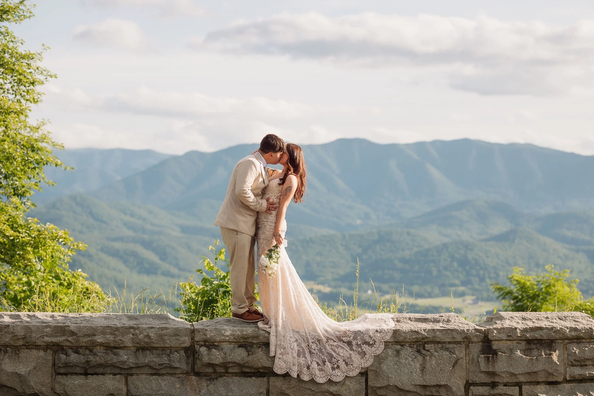 A bride and groom share a kiss at a scenic Foothills Parkway overlook, her lace wedding gown draped across the stone wall with breathtaking Smoky Mountain ridges stretching into the distance.