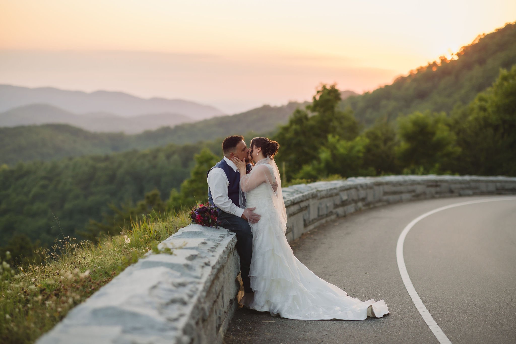 A bride and groom share a sunset kiss beside a curved stone wall overlooking layers of Smoky Mountains during their romantic Gatlinburg elopement day.