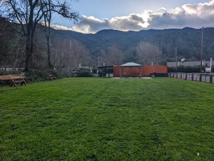 Green field with a hammock, benches, and a small wooden structure, surrounded by trees and mountains in the background under a cloudy sky