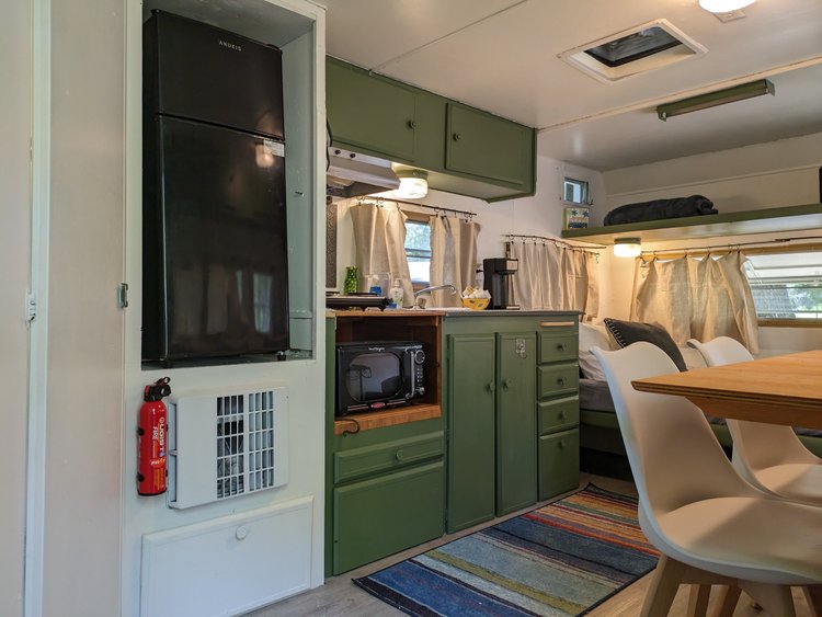 Interior of an RV featuring a green kitchenette with a black refrigerator, microwave, and coffee maker, wooden dining table, white chairs, striped