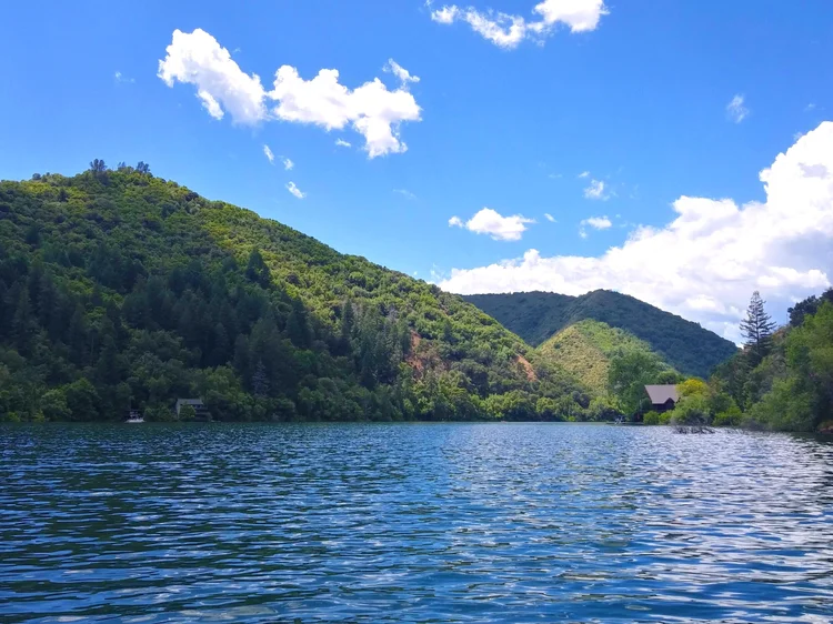 Scenic view of a lake surrounded by lush green hills and a clear blue sky, with scattered white clouds and a house by the water's edge