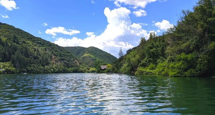 Scenic view of a lake surrounded by lush green hills and trees, with a cloudy blue sky overhead