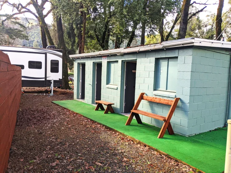 A small concrete building with doors and a window, featuring wooden benches outside, next to a white trailer in a wooded area