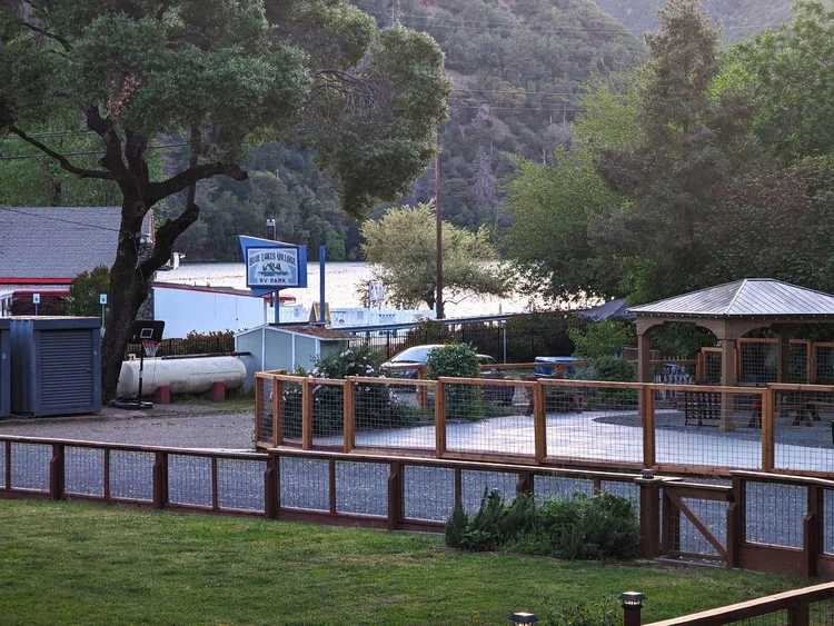 RV park entrance with a sign reading 'Blue Lakes Village RV Park', a fenced patio area, and a distant view of a lake surrounded by trees and hills