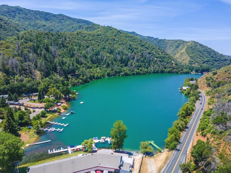 Aerial view of a lake surrounded by forested hills and a road with parked boats and docks along the shoreline