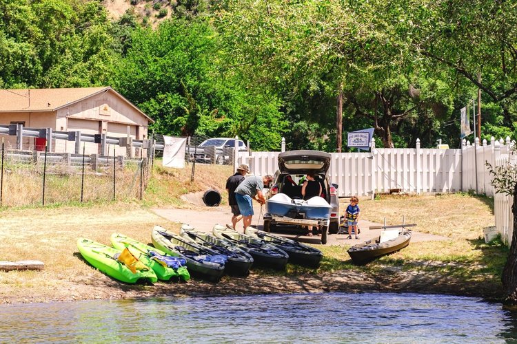 Family preparing kayaks by a lakeside, with a car parked nearby, trees and a fence in the background