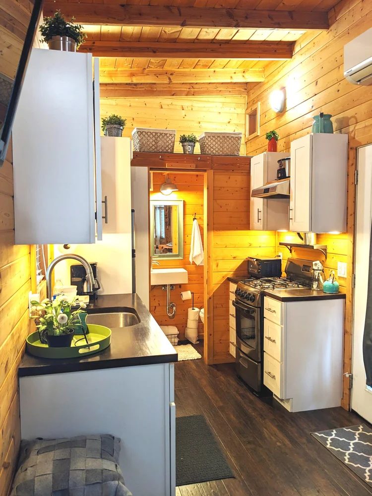 Interior of a compact wooden kitchen with white cabinets, a small sink, a stove, and decorative plants on the countertops and shelves