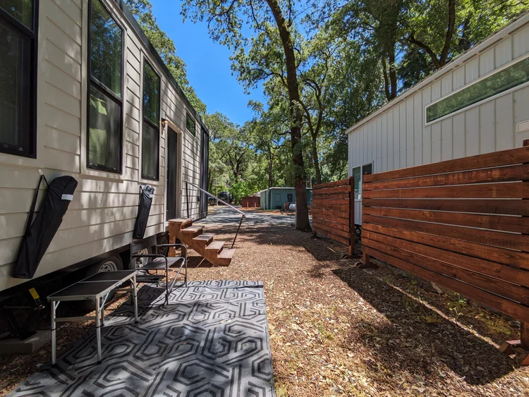 Outdoor scene with two tiny houses surrounded by trees and a wooden fence, featuring steps and camping chairs on a patio area