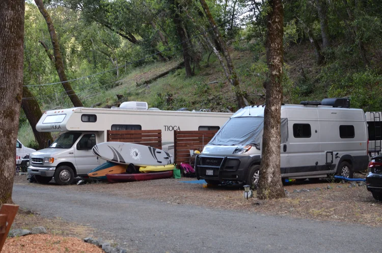 RV and camper van parked in a wooded campsite, with surfboards leaned against the vehicles