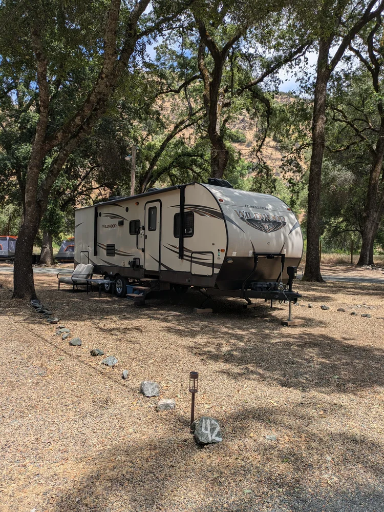 Travel trailer parked in a wooded campsite with trees and gravel, marked by rocks and a lamp with the number 42