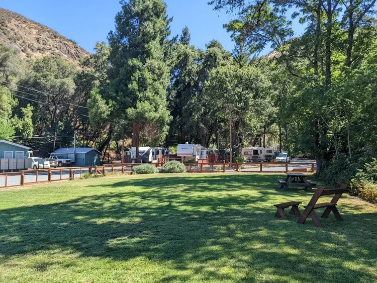 Grassy park area with picnic tables and benches, surrounded by trees, small houses, and parked trailers