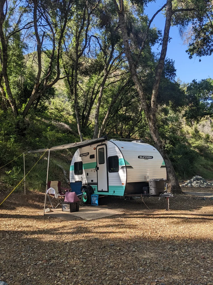 A small RV camper parked in a forested campsite with trees and blue sky