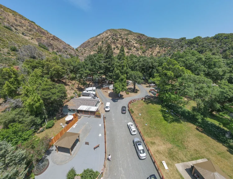 Aerial view of a scenic campground with parked cars, green trees, and surrounding hills on a sunny day