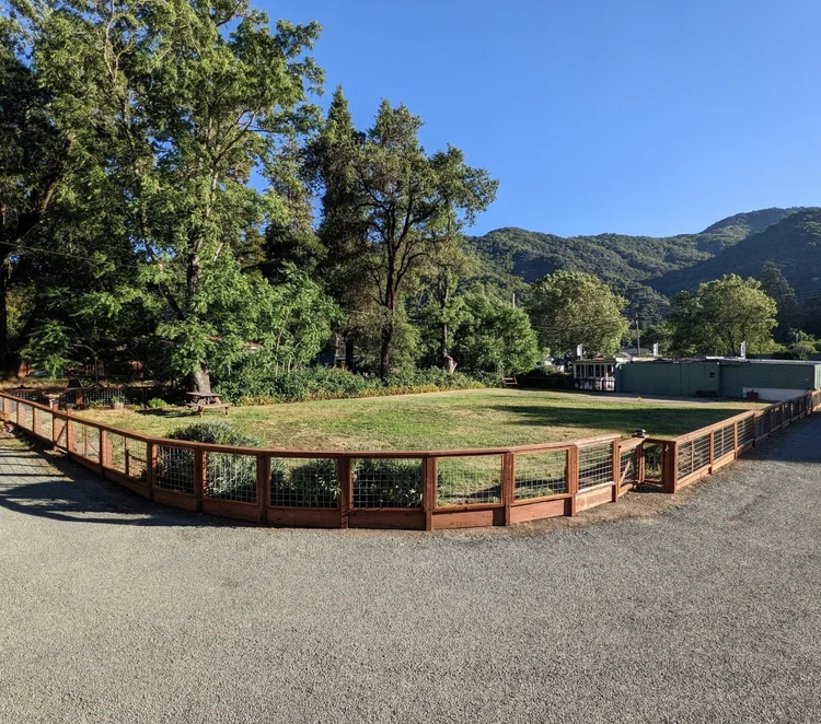 A fenced grassy area surrounded by trees and mountains under a clear blue sky