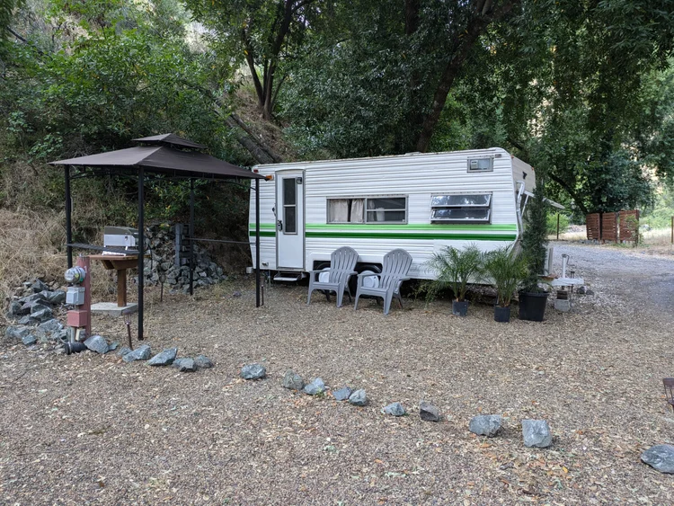 A vintage travel trailer with green accents parked in a gravel area surrounded by trees