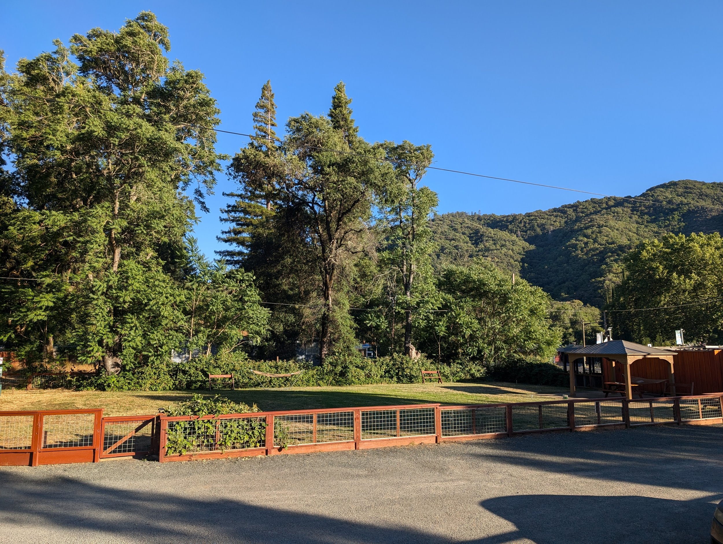 Outdoor scene with trees, grassy area, wooden structures, and mountains under a clear blue sky.