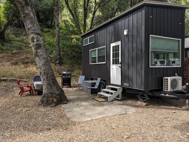 A small black tiny house on wheels with a white door and multiple windows, situated in a forested area