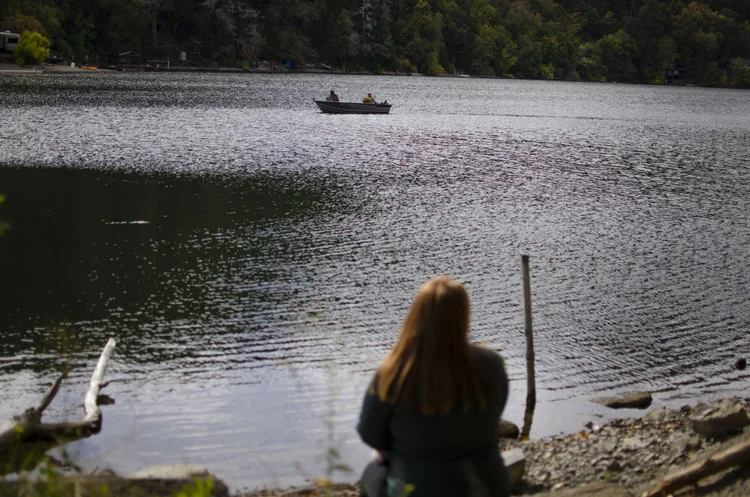 Person sitting by a lakeside watching a boat with two people on the water, surrounded by trees