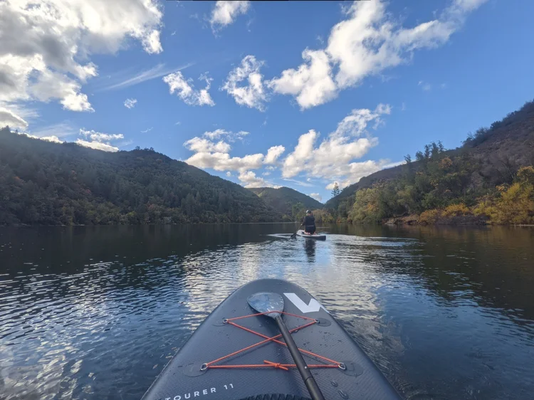 Person paddleboarding on a calm river surrounded by scenic hills and blue sky with clouds