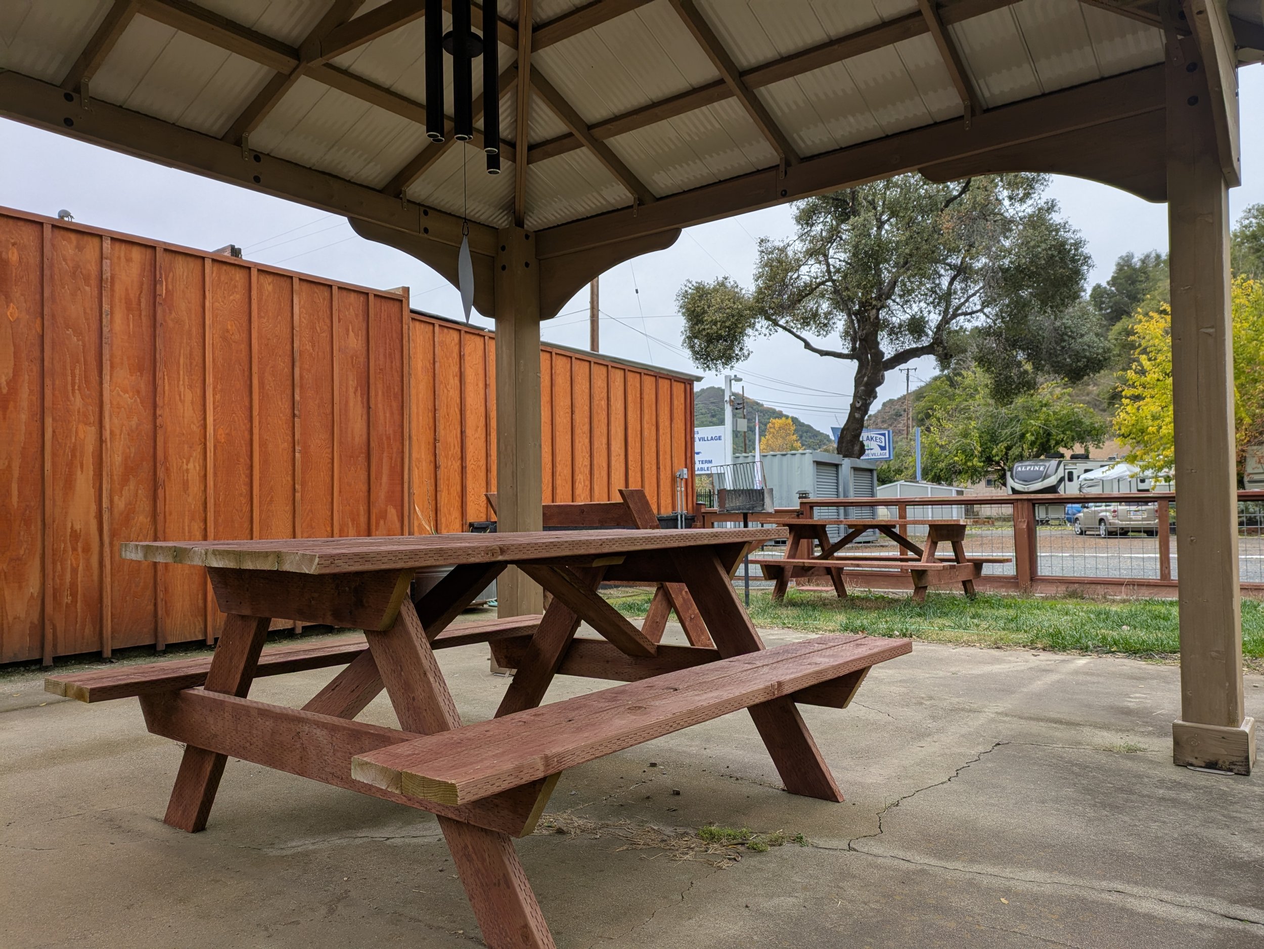 Picnic area with wooden tables under a gazebo, surrounded by a wooden fence, with trees and parked vehicles in the background.
